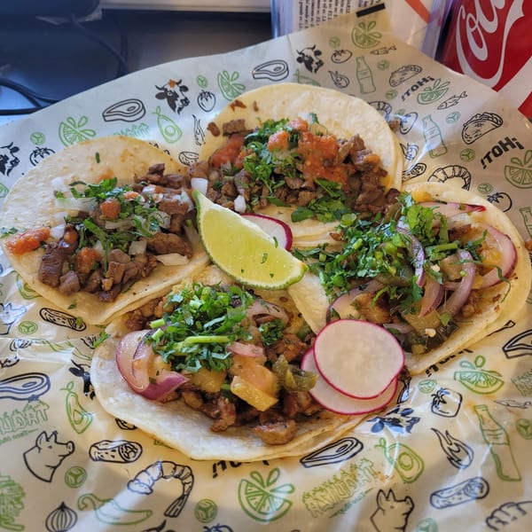 Four Taquiza Street tacos with various toppings, lime, and radishes on branded paper.