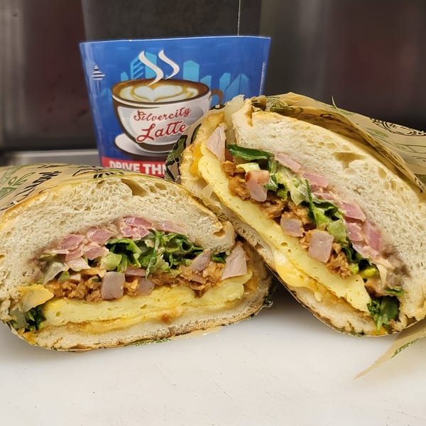 A Mexican torta sandwich, cut in half, revealing layers of meat, cheese, and vegetables, with a coffee cup in the background.