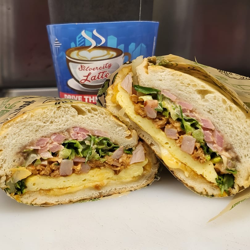 A Mexican torta sandwich, cut in half, revealing layers of meat, cheese, and vegetables, with a coffee cup in the background.