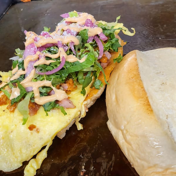 A Mexican torta with scrambled eggs, lettuce, red onions, and sauce on a griddle.