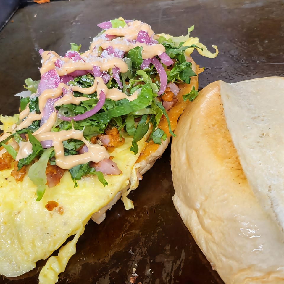 A Mexican torta with scrambled eggs, lettuce, red onions, and sauce on a griddle.