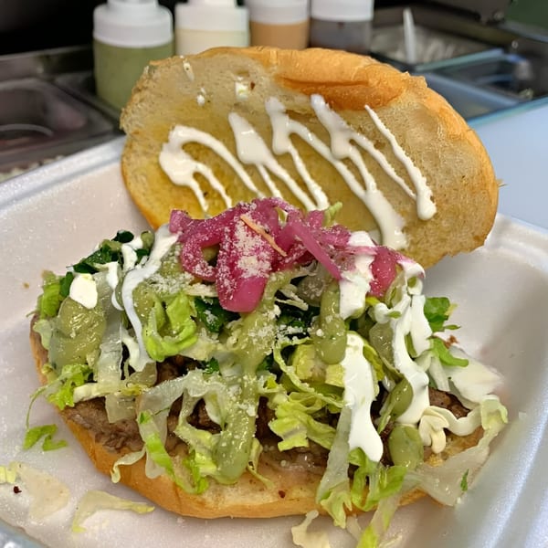 A Mexican torta with meat, lettuce, pickled onions, and various sauces on a white tray.