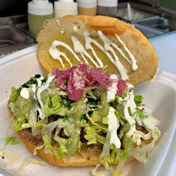 A Mexican torta sandwich, overflowing with meat, lettuce, pickled onions, and various sauces in a styrofoam container.
