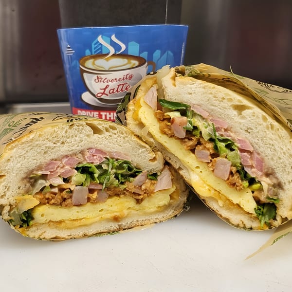 A large Mexican torta sandwich, cut in half, revealing layers of egg, meat, and lettuce, with a coffee cup in the background.